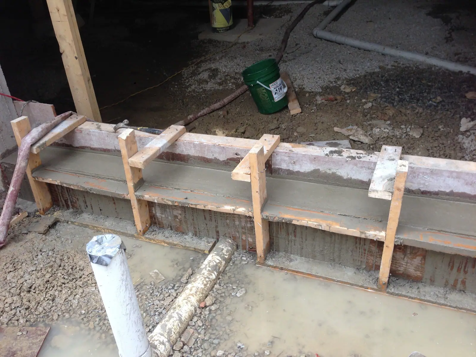 A form of boards and stakes along a poured edge, with a white drain pipe standing in the gravel beside it.