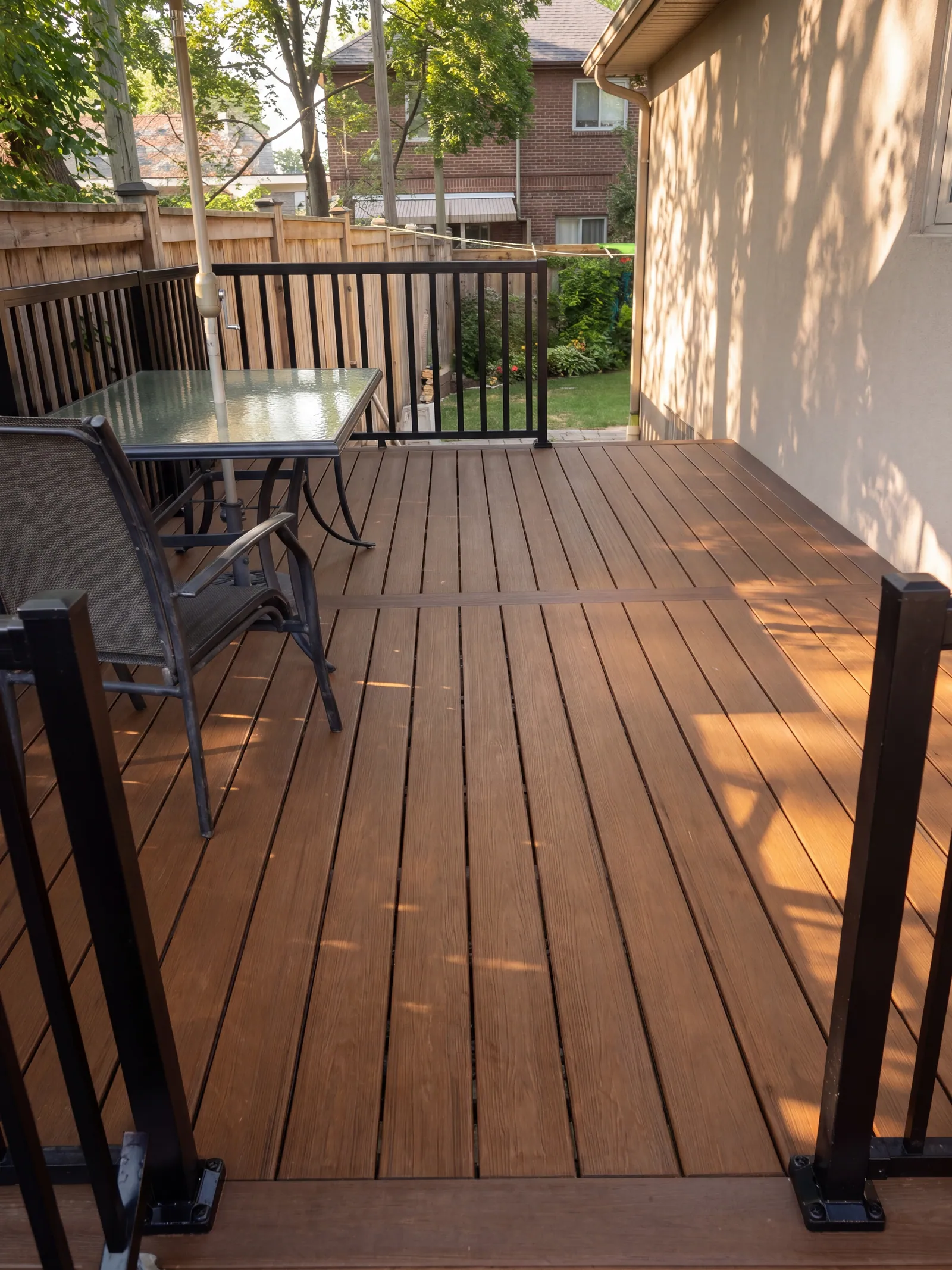 A stained deck with a black metal railing, a glass-topped patio table and chairs, and a green yard with neighbouring houses beyond.