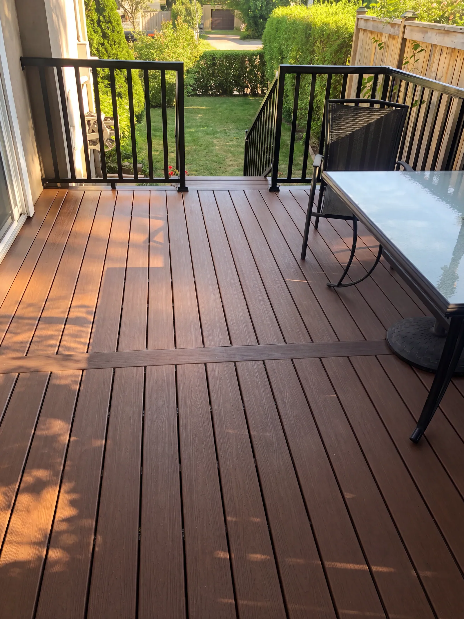 Stained deck boards in low sun with a black metal railing, a chair and a glass-topped table, and a green hedge beyond.