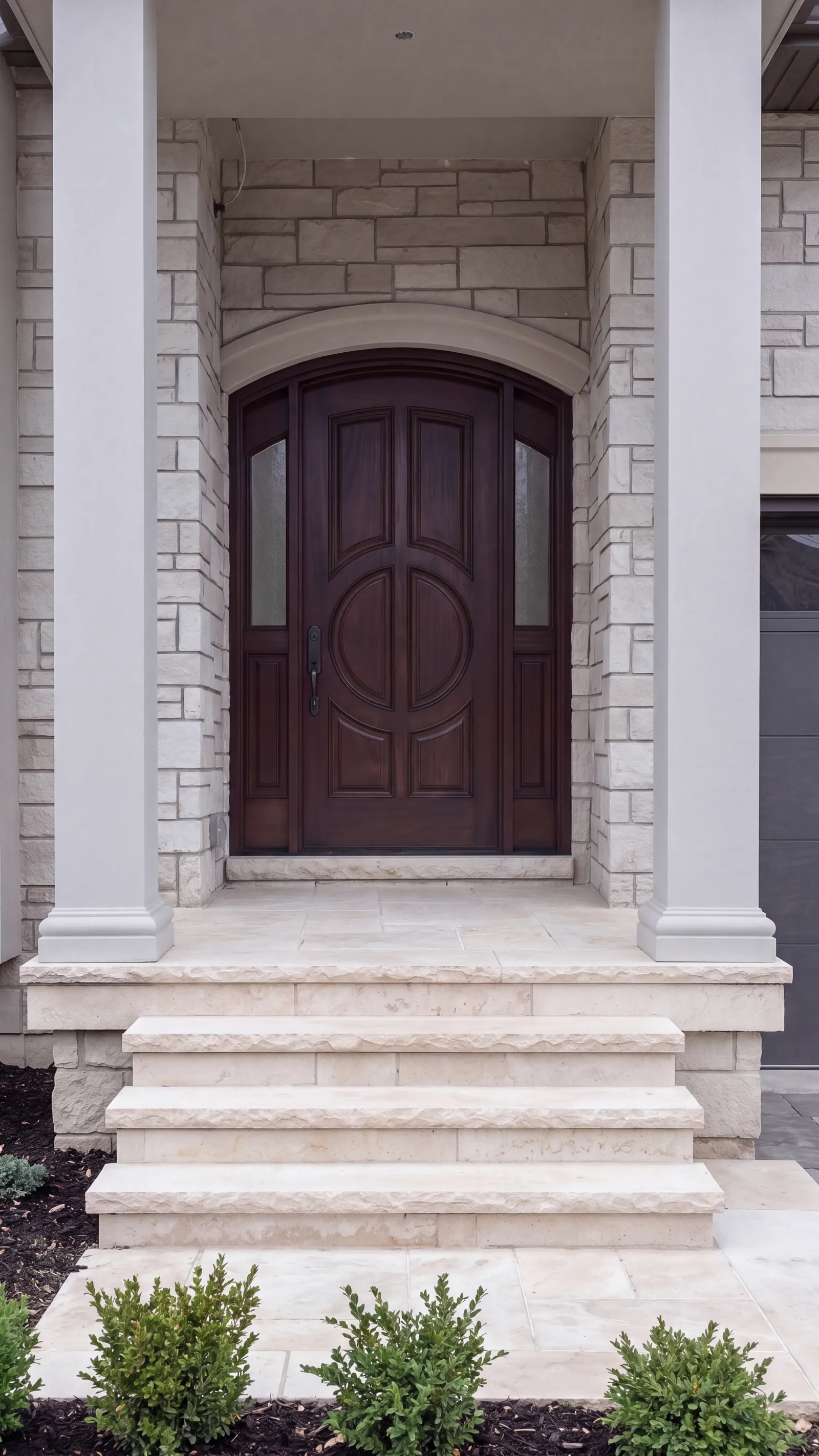 An arched dark wood front door with glass sidelights in a stone surround, between two square white porch columns, with stone steps and shrubs in front.