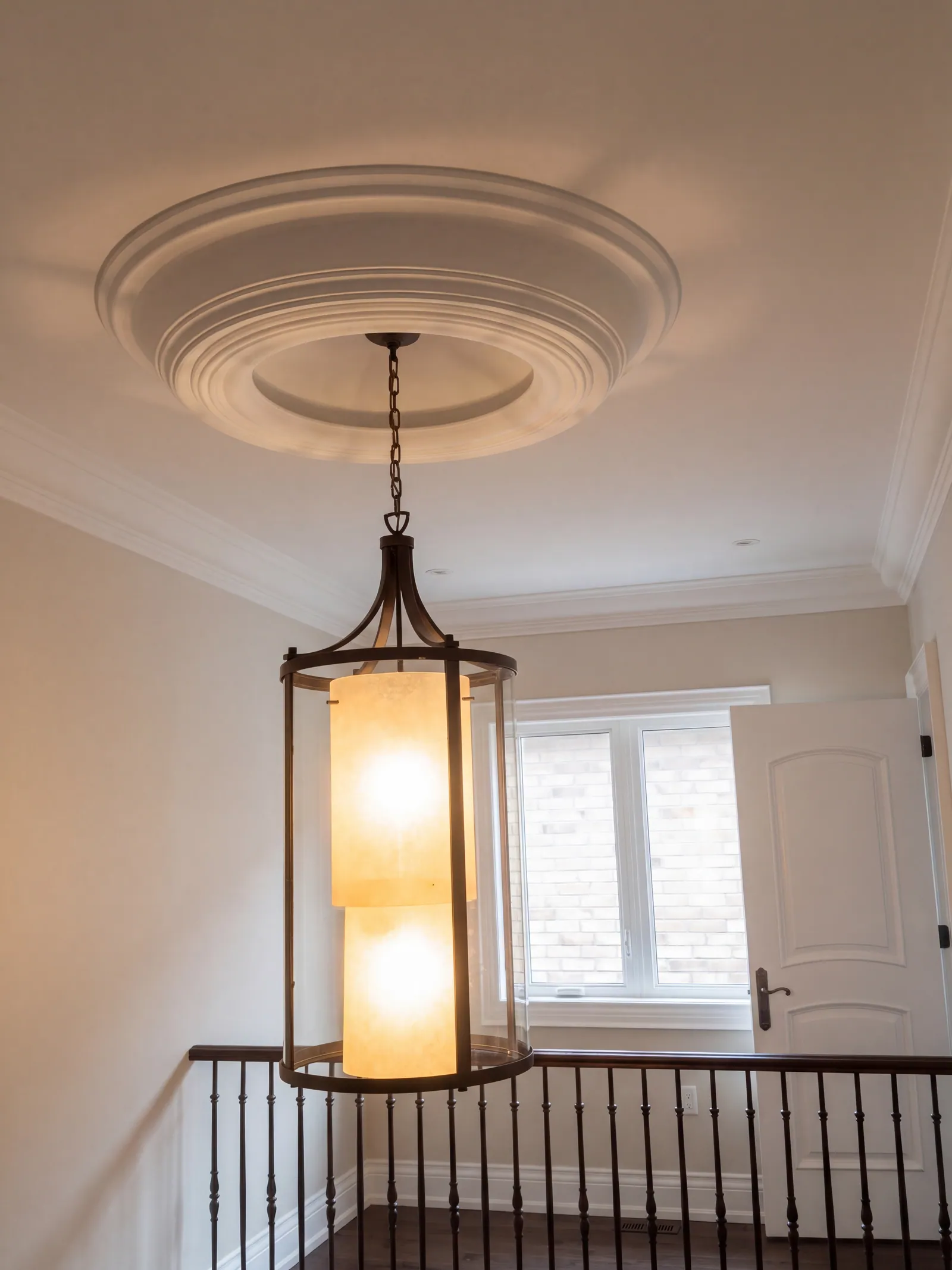 A lit lantern-style pendant light under a round ceiling medallion, with a window and a door beyond and a black stair railing below.