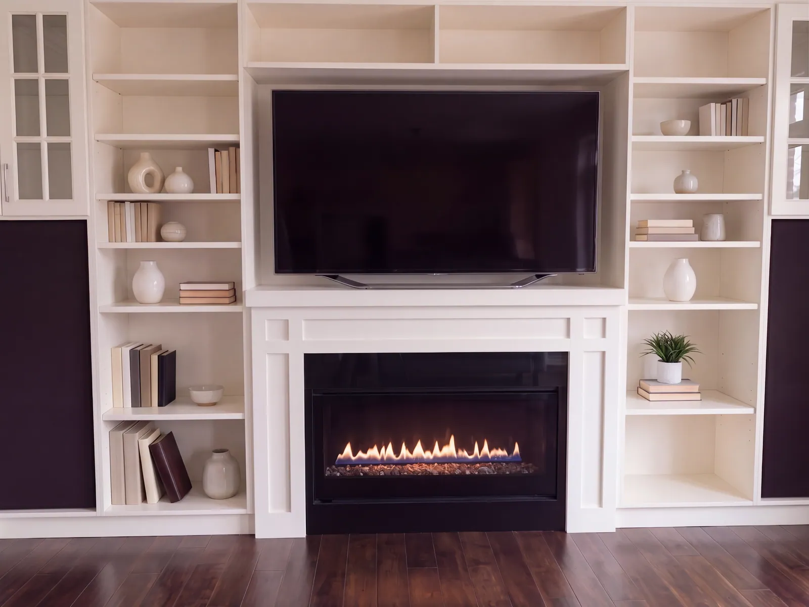 A white built-in wall of shelves and cabinets around a wall-mounted television and a lit gas fireplace, with books and vases on the shelves, over a dark wood floor.