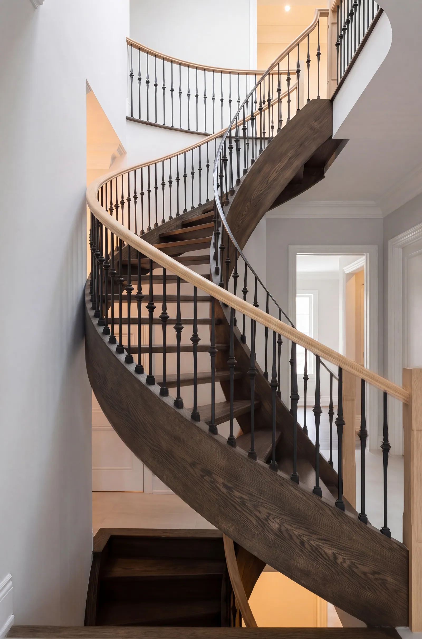 A curved open-tread stair rising through a foyer, with a dark stained curved stringer, black iron balusters, a pale wood handrail and a pale wood newel post at the right.