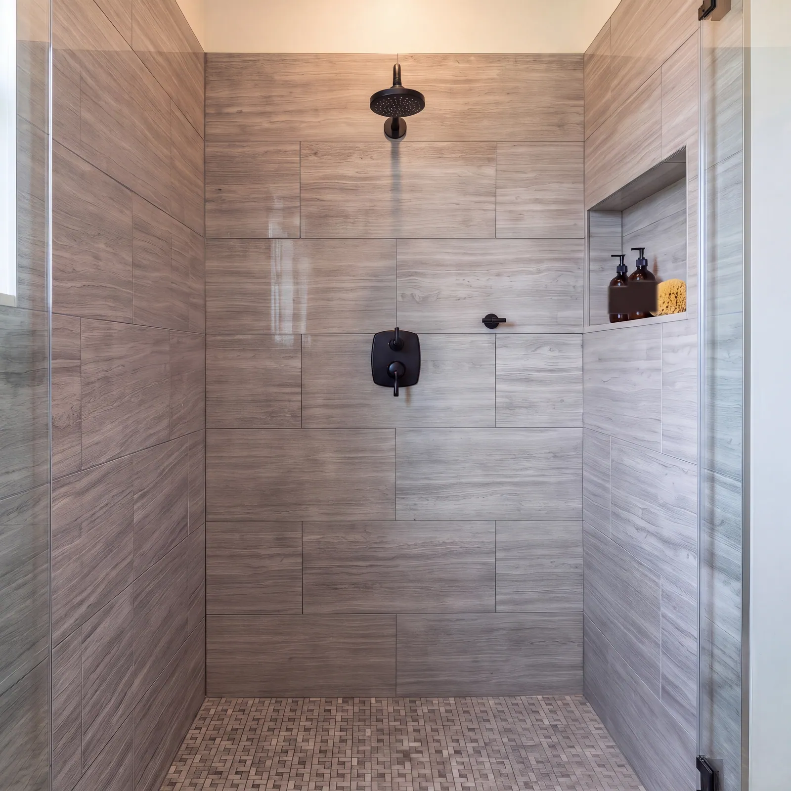 A walk-in shower with grey wood-grain tiles on the walls, a mosaic floor, a black shower valve, a black shower head and arm, a recessed niche holding two amber bottles and a sponge, and a glass door.