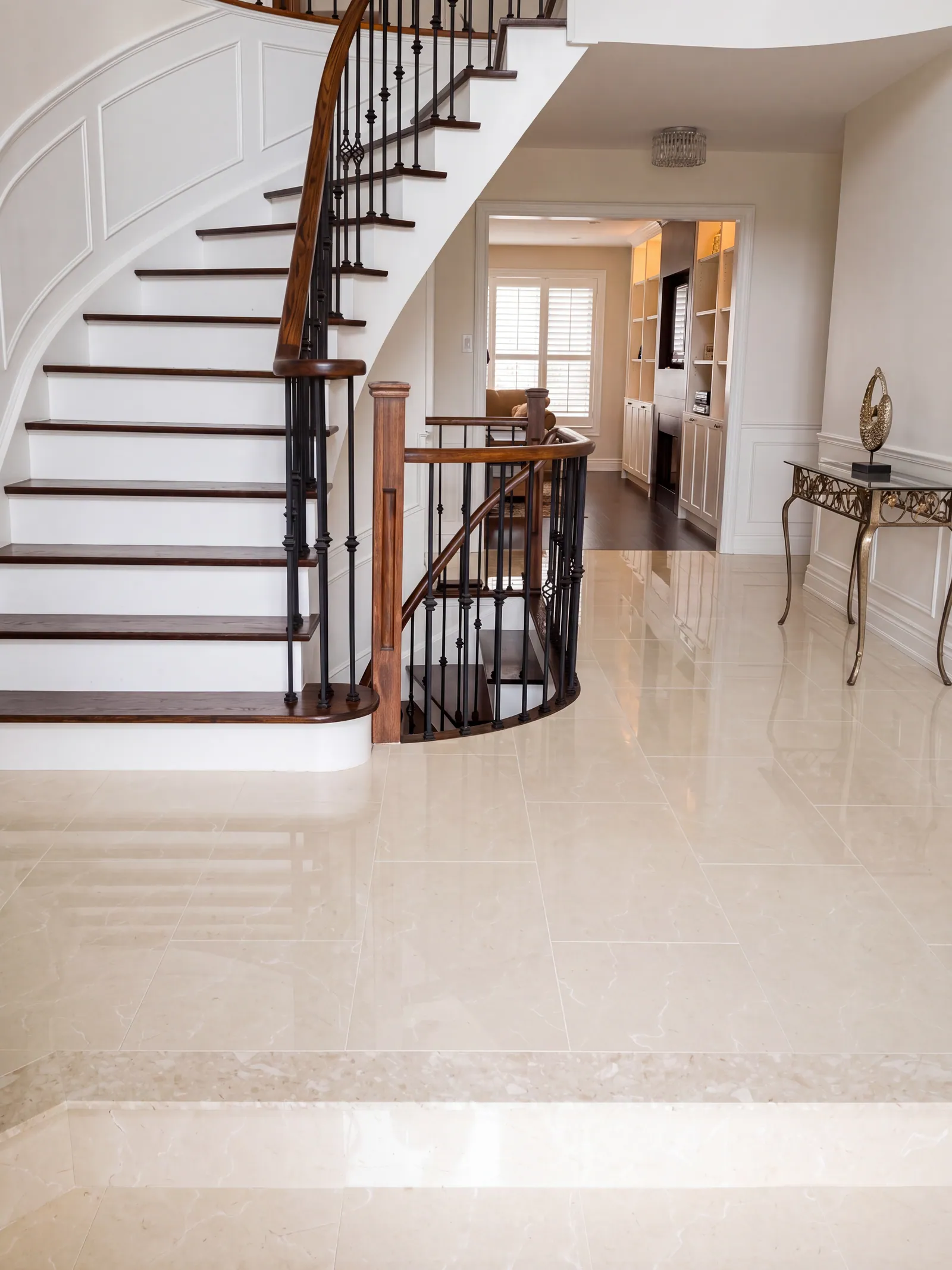 A curved stair with white risers and dark wood treads beside a polished cream stone foyer floor, with a console table against the wall and a view through to a kitchen.