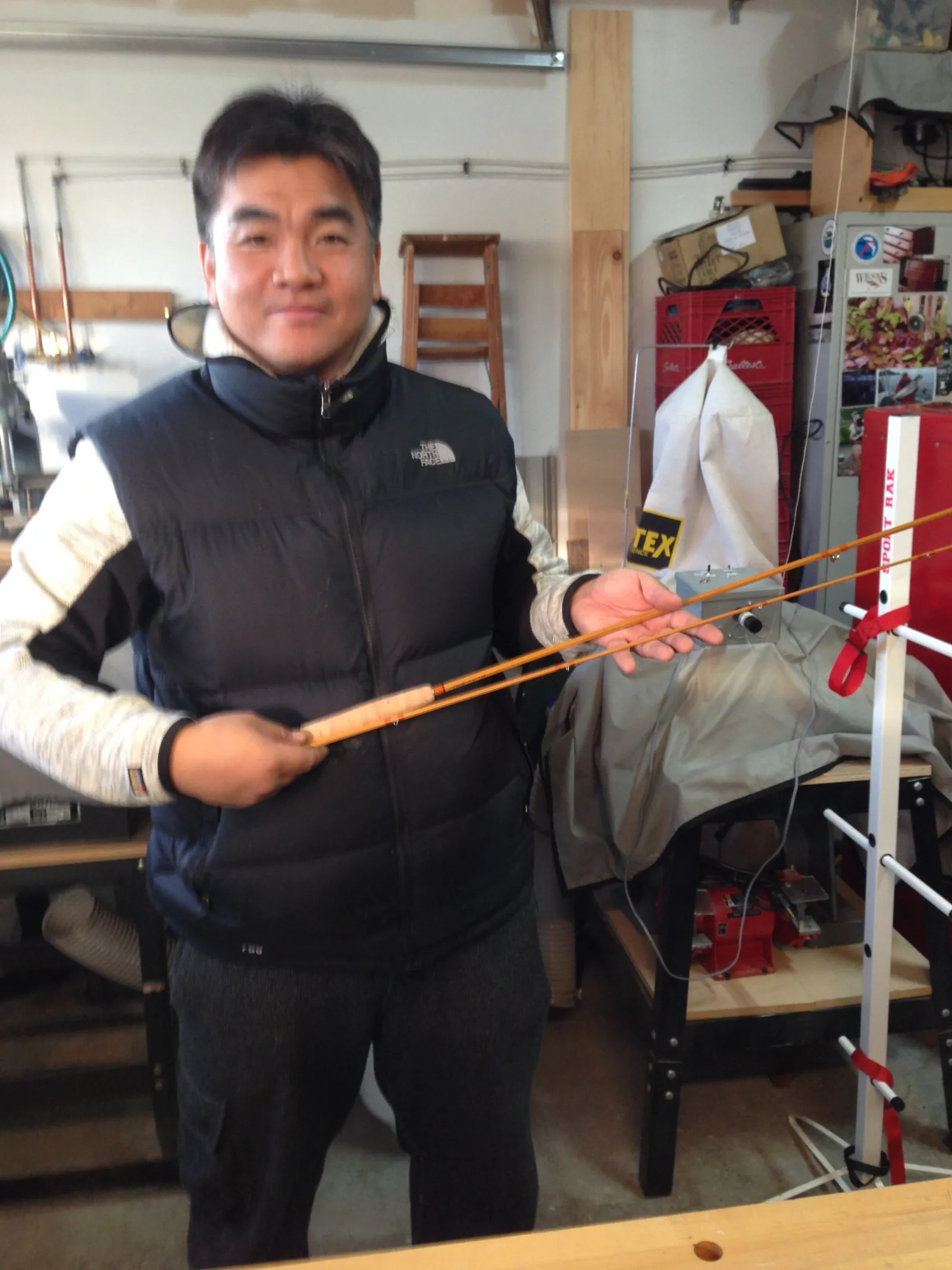 Young in a black vest holding a bamboo rod in a workshop.