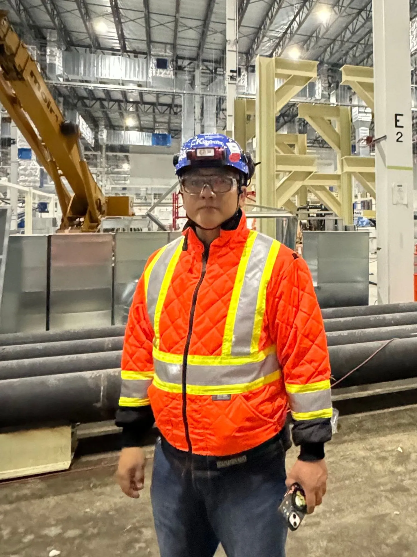 Young in an orange hi-vis jacket and a blue hard hat, standing on a large indoor industrial site.